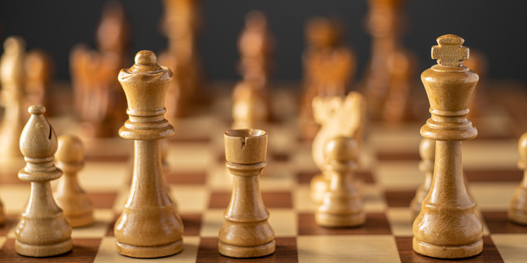 Chess pieces set up on a chess board in the middle of a game during the opening phase. The white pieces are nearest us with the black pieces out of focus in the background. There is left to right a bishop, queen, rook and king on the nearest row